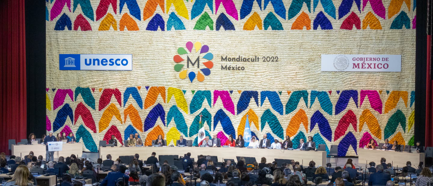 Publik som sitter framför scenen. Scenen har en vägg fylld med loggor och färgglada illustrationer. 