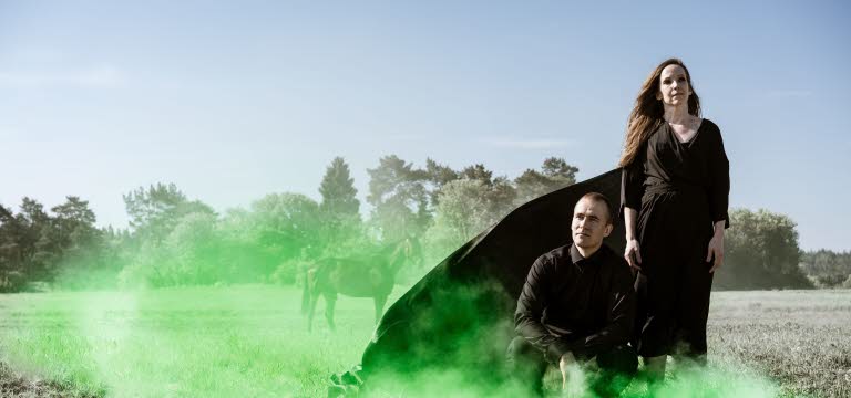 En man och en kvinna i svart klädsel på ett torrt gräsfält med grön rök.