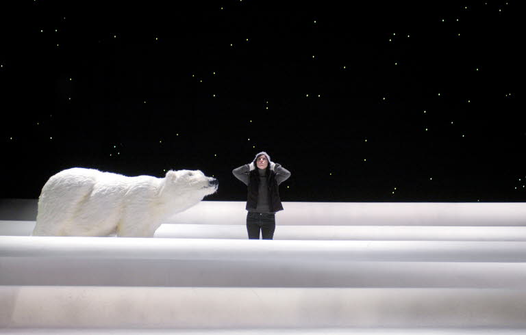 A girl stands on a stage, the background is black and she has snow to her knees, on her right is a polar bear. 