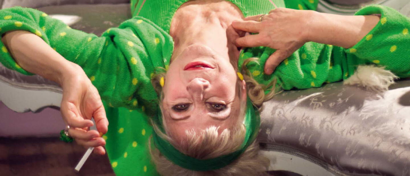 Book cover of "Vem tror hon att hon är": it's a photo of a women lies upside down on a couch wearing a green sweatsuit with green dots, smoking a cigarette.
