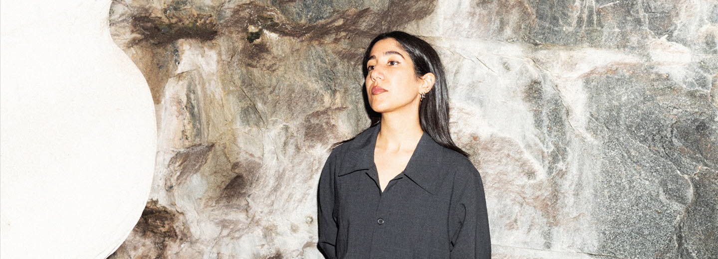 Leila is wearing black clothes and leaning to a stone wall.