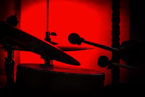 Trumma, cymbaler och trumstockar i silhuett mot rödbelyst bakgrund.