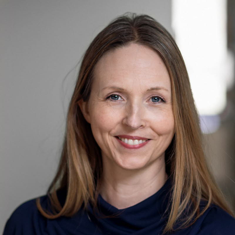 Medelålders kvinna med långt cendréfärgat hår och blå tröja omgiven av bokhyllor.