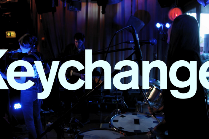 Vit text med Keychange, i bakgrunden ett band som spelar.