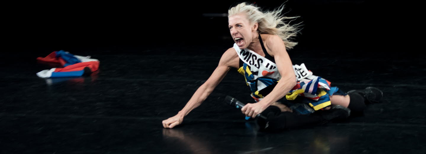 En kvinna med mikrofon halvliggandes på ett scengolv.