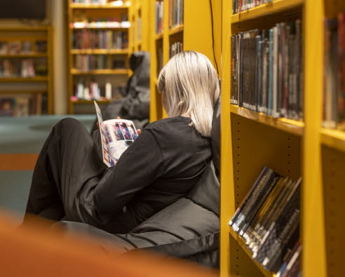 Tonårstjej sitter i fåtölj på bibliotek och läser en bok.