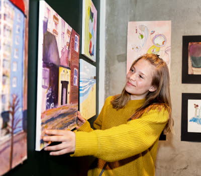 Ett barn rättar till en tavla i ett rum fyllt av målningar. Hon ser glad ut.