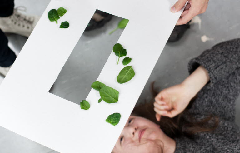 En kvinna ligger på rygg och tittar upp mot ett genombrutet papperark som hålls ovanför hennes huvud. Det ligger gröna blad på pappersarket. 