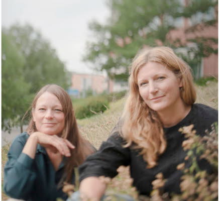 Foto på Jenny och Lotta som sitter ner i gräset, i bakgrunden skymtas träd och en tegelbyggnad.