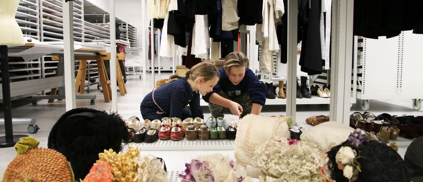 Två barn, pojke och flicka, i museums magasin pekar på samling skor.