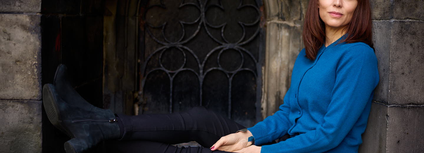 Lina Wolff sits in a window with her legs up, she is looking into the camera and wearing a blue shirt.