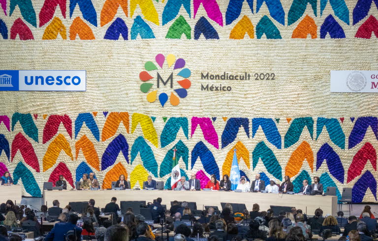 Publik som sitter framför scenen. Scenen har en vägg fylld med loggor och färgglada illustrationer. 