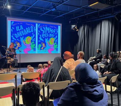 Två personer sitter på en scen och läser framför en publik med barn och vuxna.