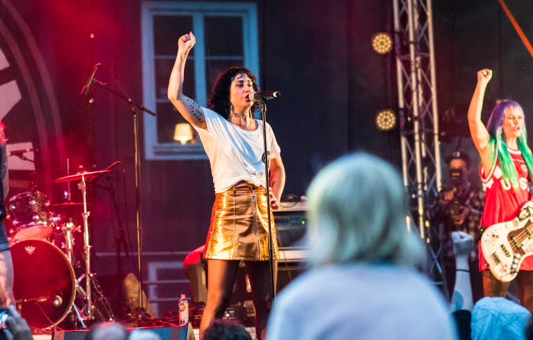 Three performers on a music stage raising their right hand and singing.