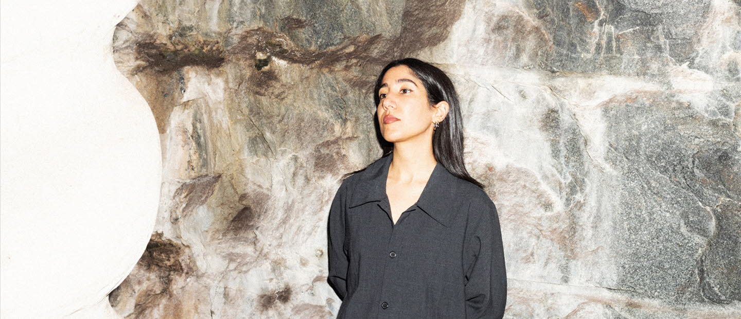 Leila is wearing black clothes and leaning to a stone wall.