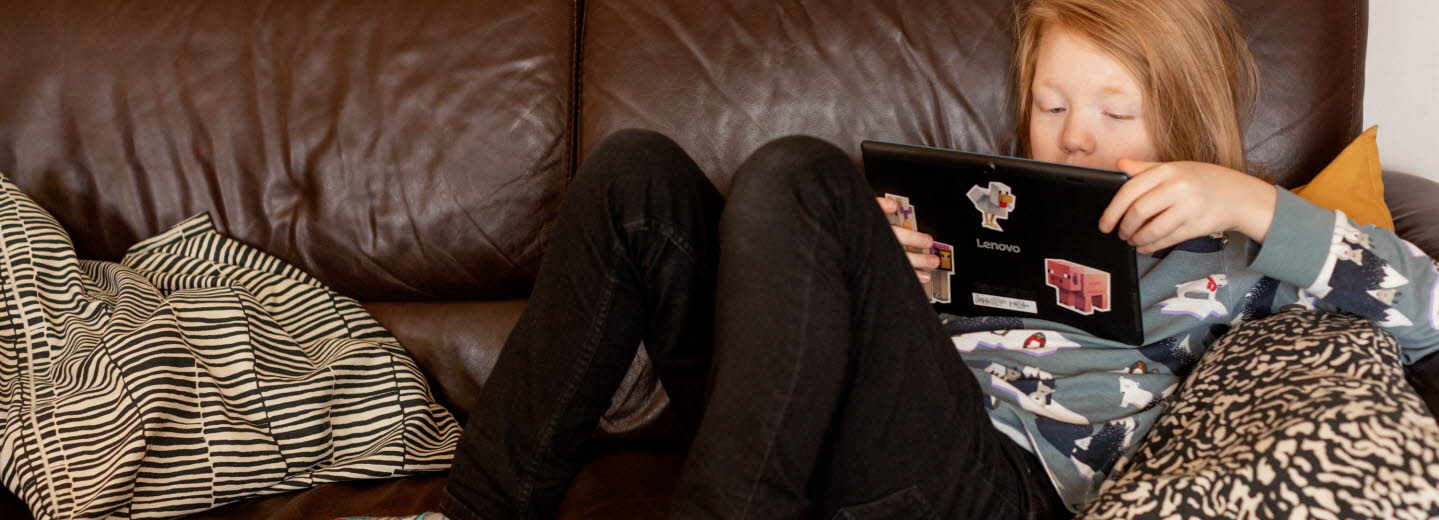 Pojke ligger i brun soffa och läser på sin Ipad. 