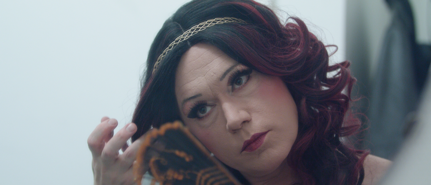 Närbild på uppsminkad dragqueen som håller i en solfjäder. Långt violettfärgat hår och guldigt  diadem.