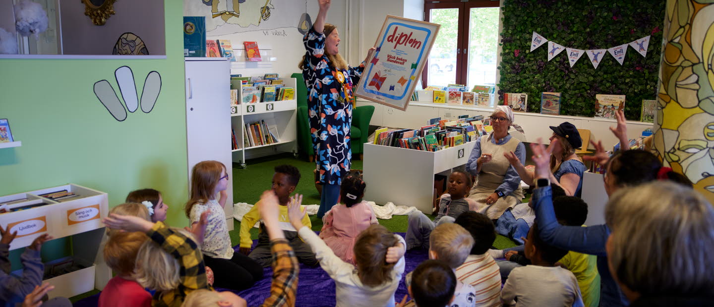 Barn som sitter i en halvcirkel kring en kvinna som håller upp ett stort diplom. 