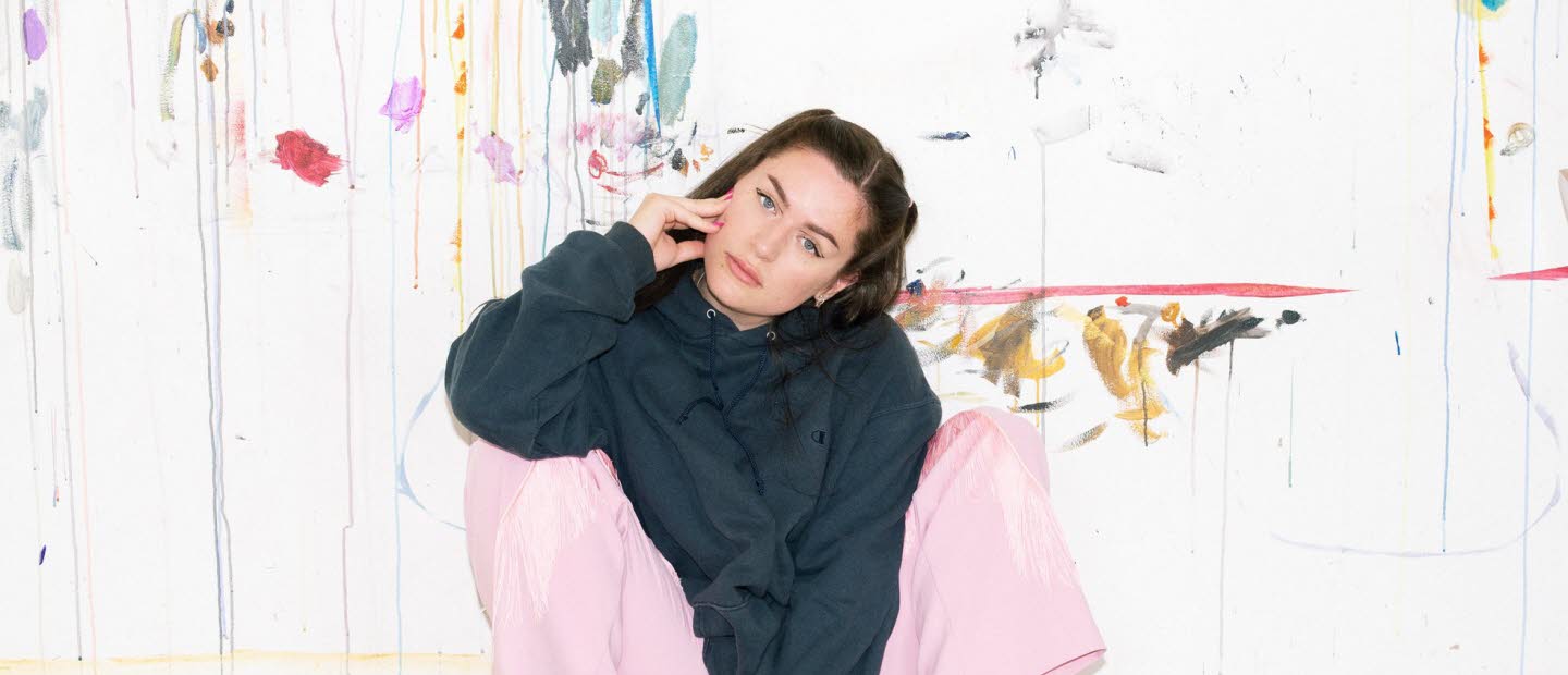 Artist Moa Romanova is sitting against a wall in pink pants and a black hoodie. 