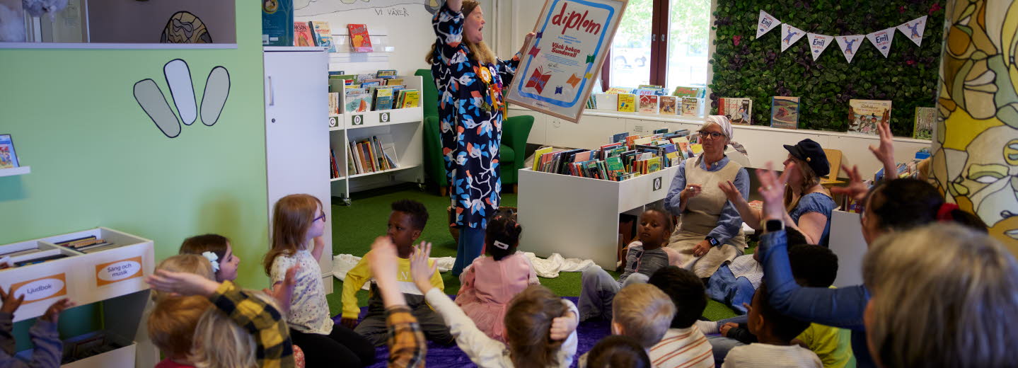 Barn som sitter i en halvcirkel kring en kvinna som håller upp ett stort diplom. 