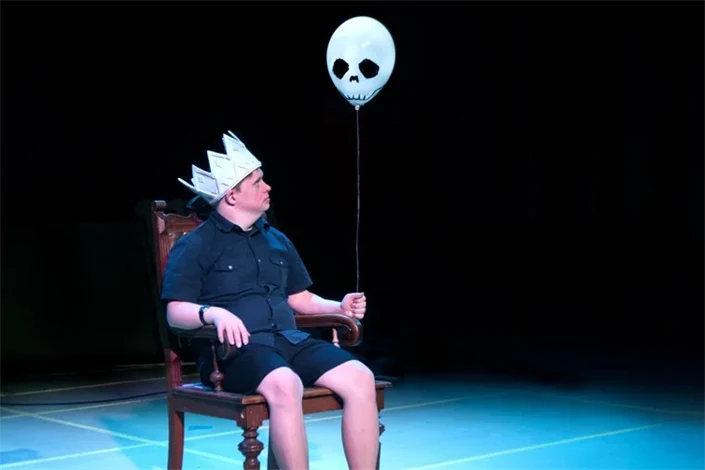 En man sitter på en scen. På huvudet har han en kungakrona i papp, i handen håller han en ballong med spökansikte.