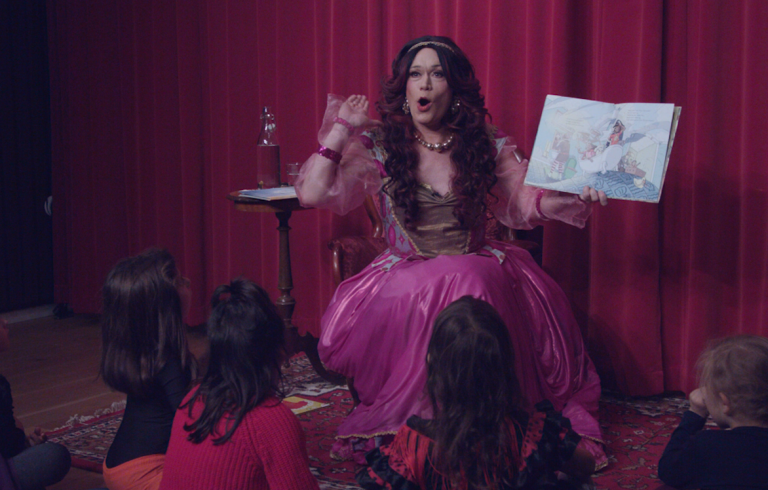 En dragqueen som läser högt för en grupp barn som sitter på golvet. 