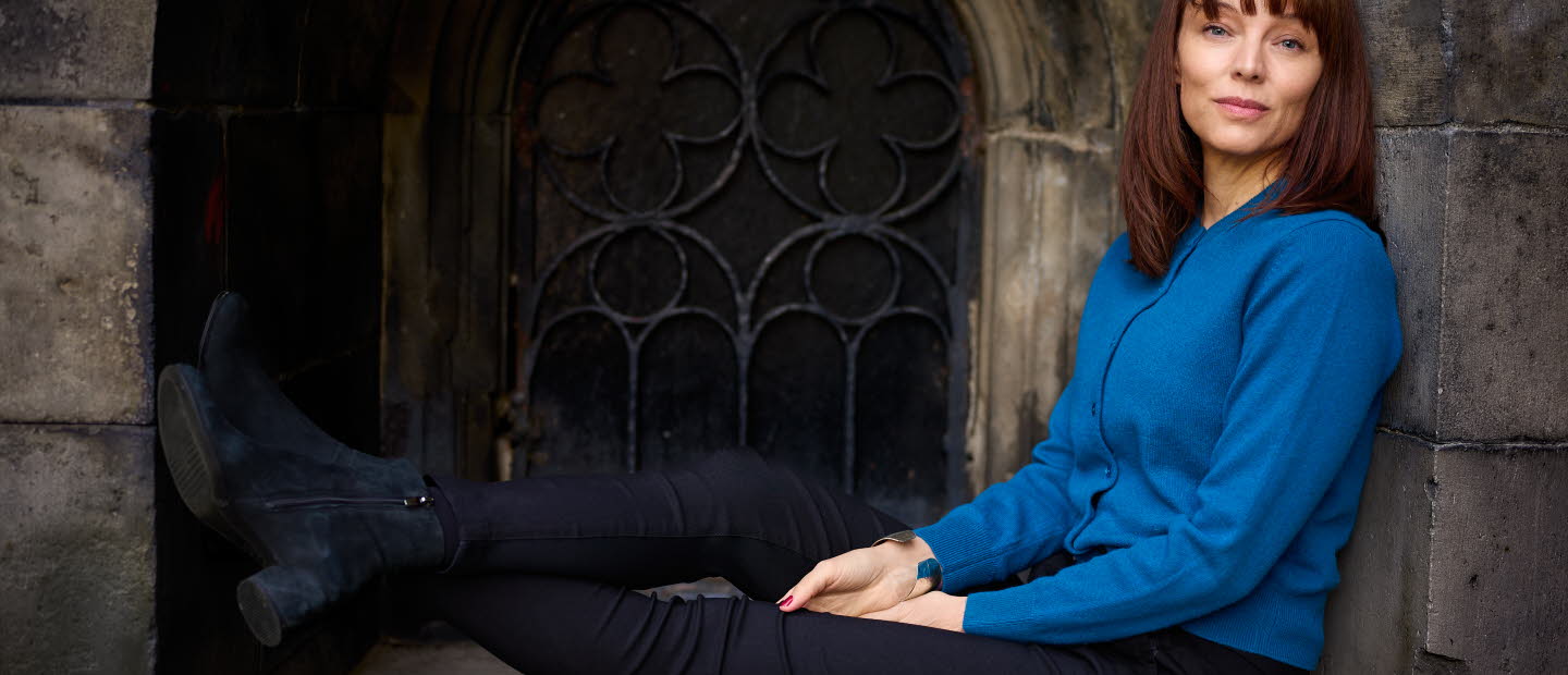 Lina Wolff sits in a window with her legs up, she is looking into the camera and wearing a blue shirt.