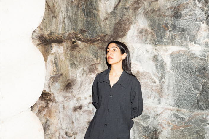 Leila is wearing black clothes and leaning to a stone wall.