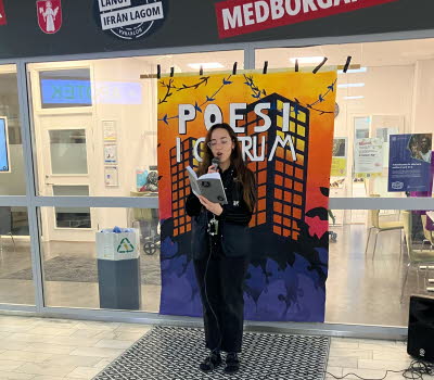 En ung kvinna står med en mikrofon i ett köpcentrum och läser ur en bok.