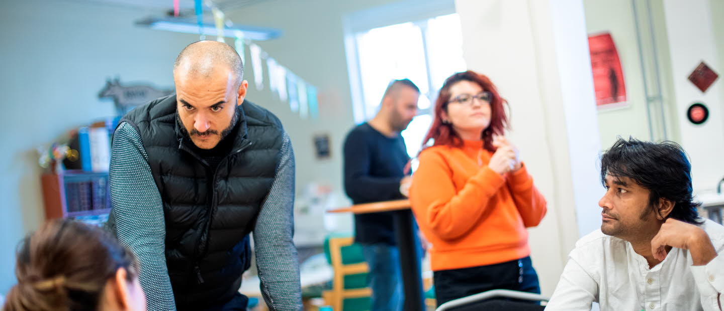 Fem vuxna rör sig i ett rum som påminner om ett klassrum. En man står upp och tittar på två sittande personer. Det ser ut att vara en kurs för vuxna.