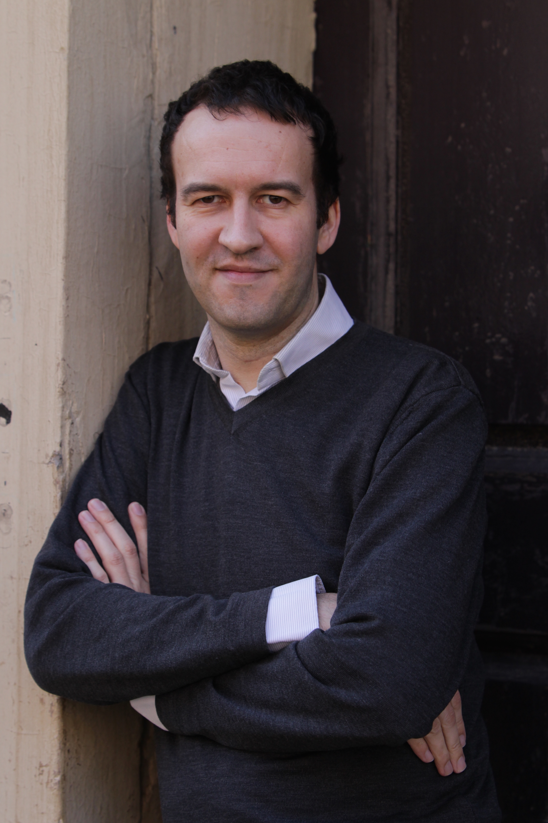 Martin Colthorpe leans against the wall and looks into the camera. He is wearing a dark jumper with a light-coloured shirt underneath.