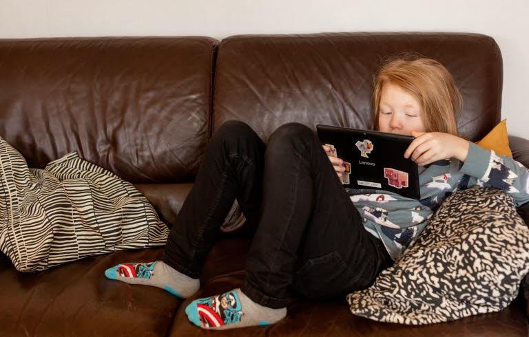 Pojke ligger i brun soffa och läser på sin Ipad. 