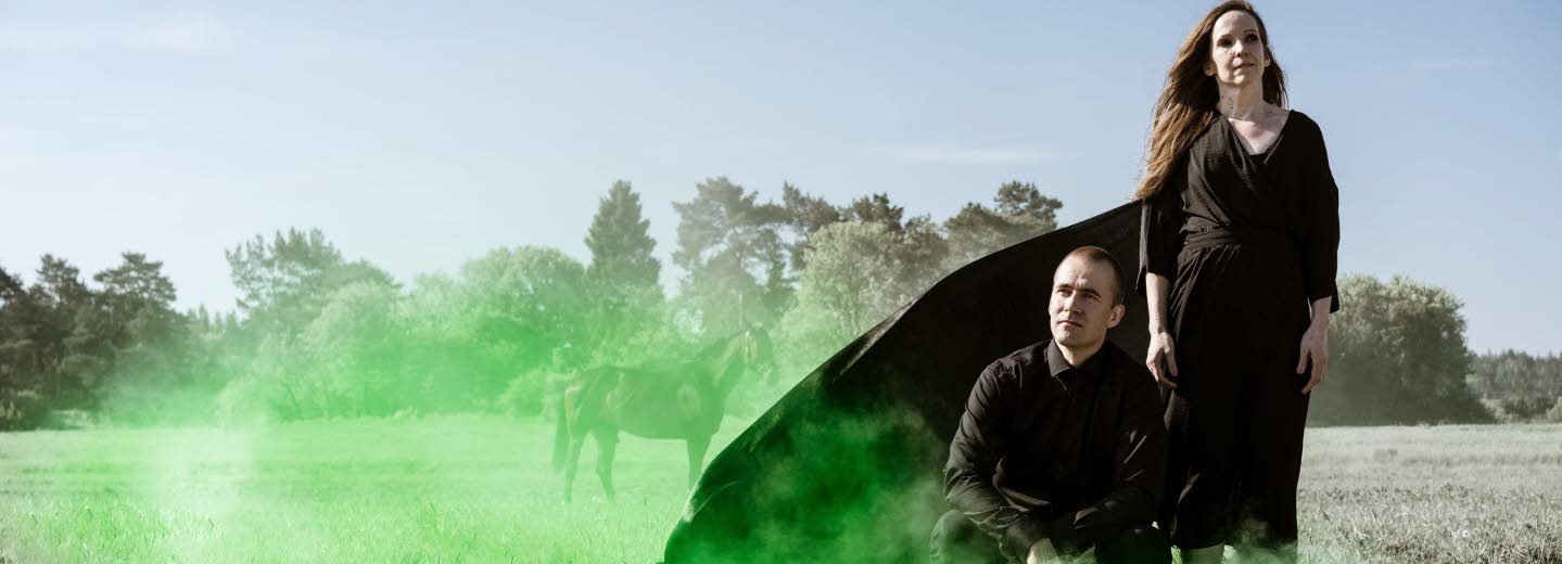 A Radar Styled Novel. Foto: Mats Bäcker. En man och en kvinna i svart klädsel på ett torrt gräsfält med grön rök.