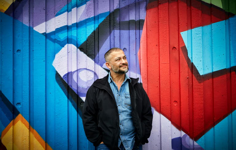 Bagir Kwiek står framför en vägg med graffiti och ler. Han tittar snett ut ur bild. 