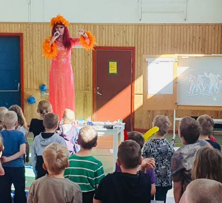Dragqueen artisten Inga Tvivel står på en scen och undervisar en skolklass. 