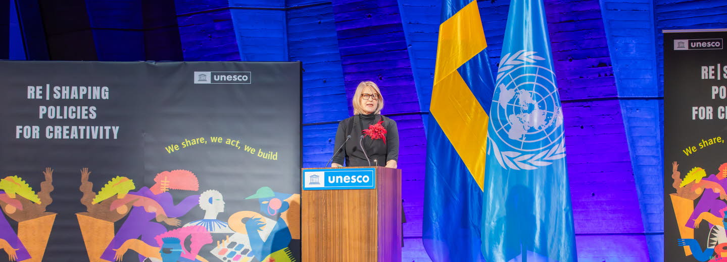 Kvinna som talar för en sittande publik. Unescos textlogotyp på talarstolen.