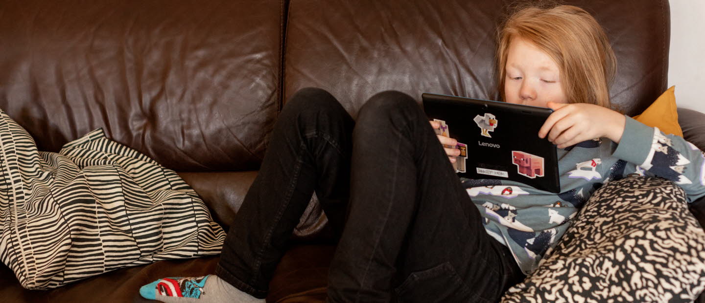 Pojke ligger i brun soffa och läser på sin Ipad. 