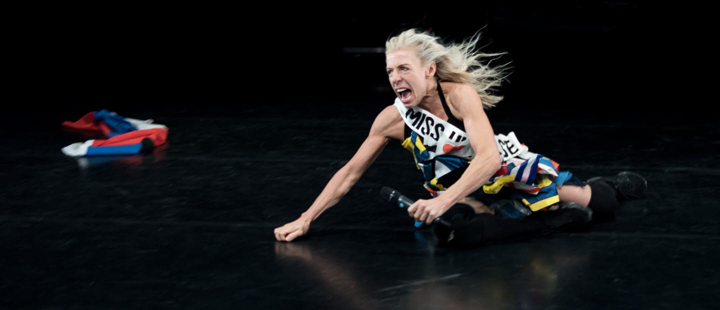 En kvinna med mikrofon halvliggandes på ett scengolv.