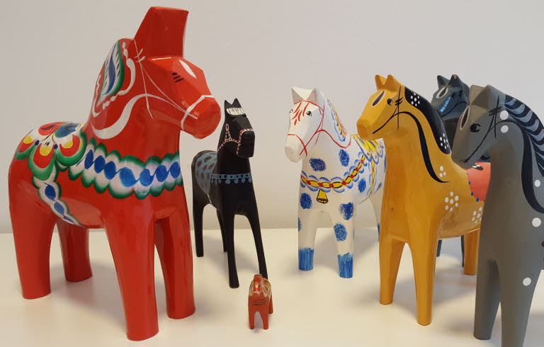 Flera dalahästar i olika storlekar och färger stående på ett vitt underlag.