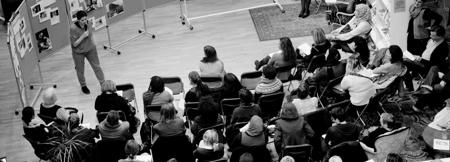  Föreläsning om Women Making HERstory föreläsningssal. Ett 30-tal människor lyssnar till en som står och pratar. Bilden är tagen uppifrån och man ser ingens ansikte tydligt.