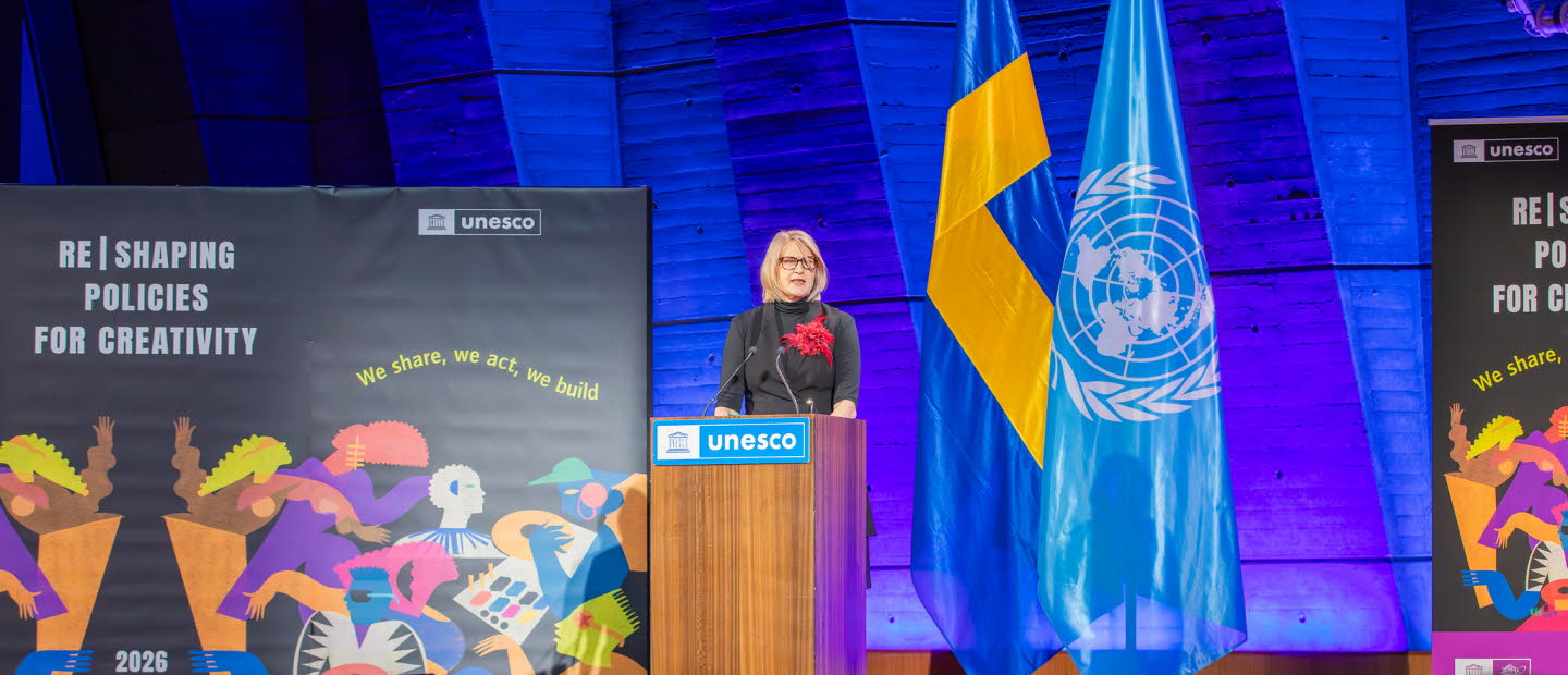 Kvinna som talar för en sittande publik. Unescos textlogotyp på talarstolen.