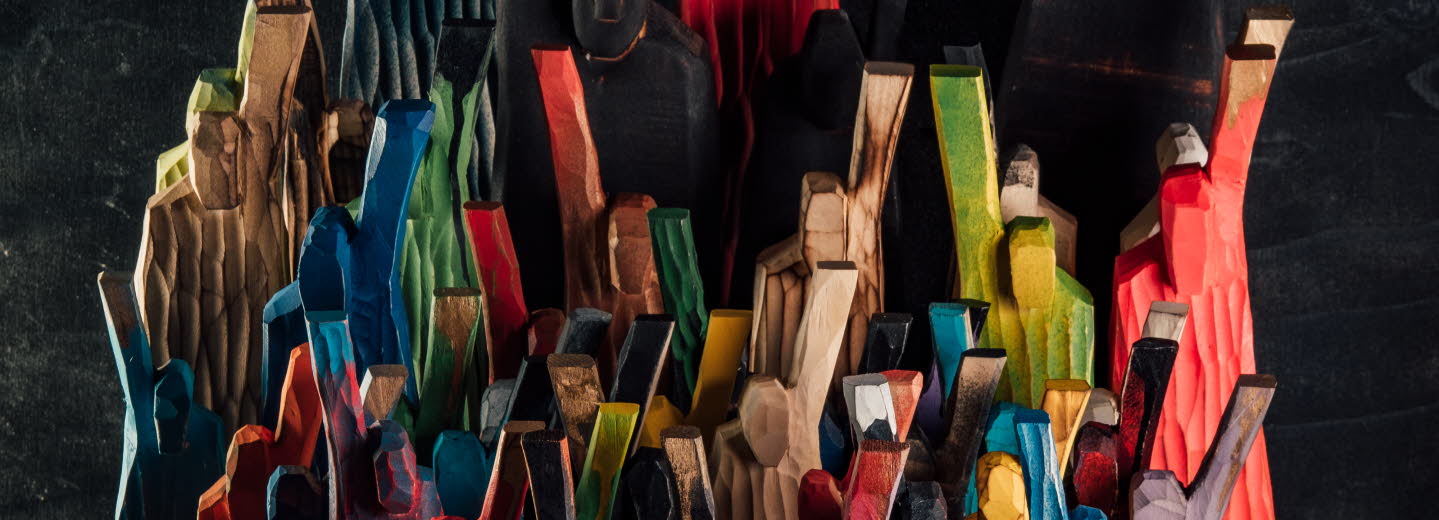 Flera färgglada, grovt täljda träfigurer står tätt tillsammans mot en mörk bakgrund, med armar lyfta i olika vinklar.