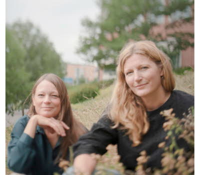 Foto på Jenny och Lotta som sitter ner i gräset, i bakgrunden skymtas träd och en tegelbyggnad.