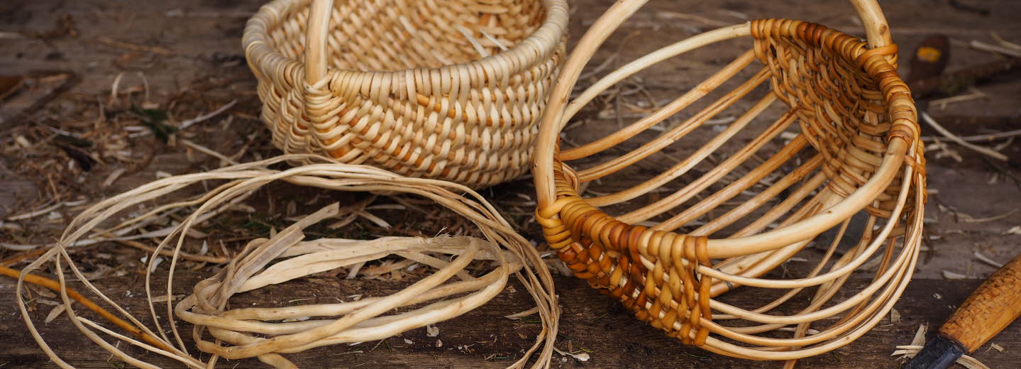Två flätade korgar i naturmaterial, omgivna av rötter och en kniv på ett träbord.
