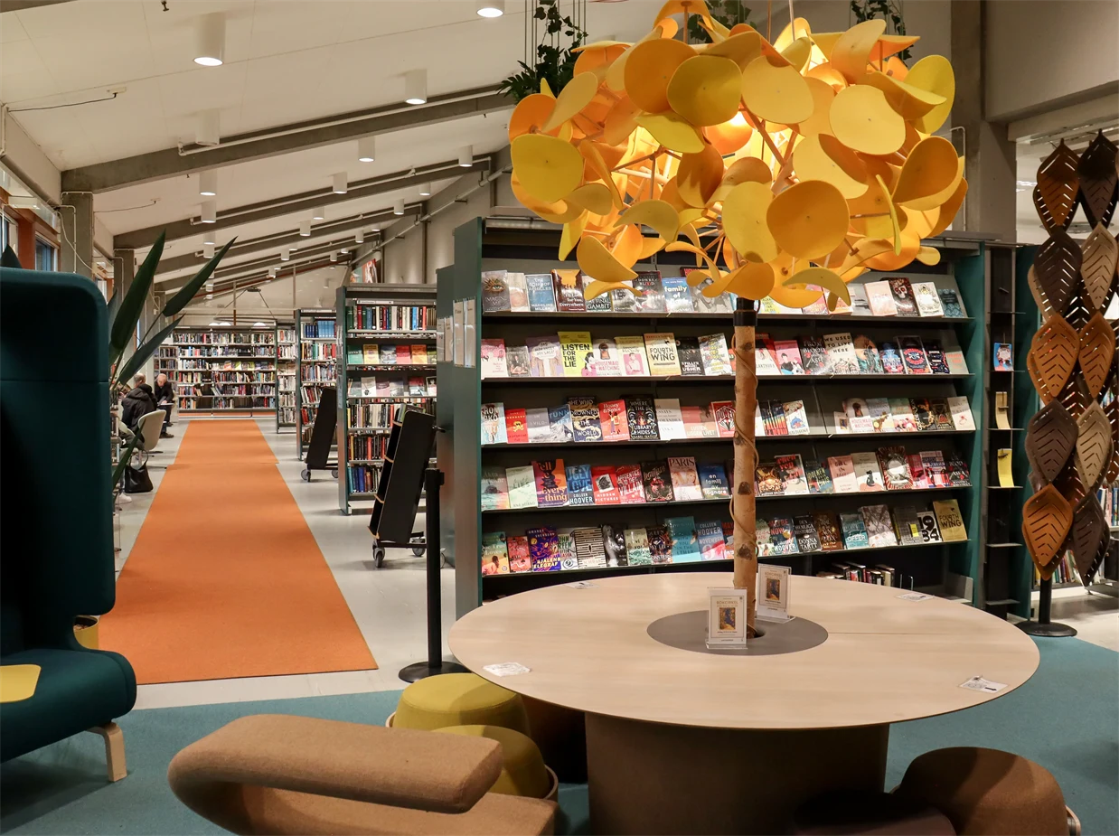 Interiör från bibliotek med bokhyllor, sittmöbler och samlingsbord med en lampa som ser ut som ett träd.