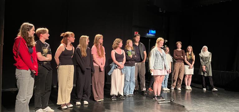En rad ungdomar står på en scen, en ung kvinna står lite längre fram vid en mikrofon.