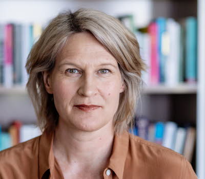 Medelålderskvinna i halvfigur. I bakgrunden bokhyllor fyllda med böcker. 