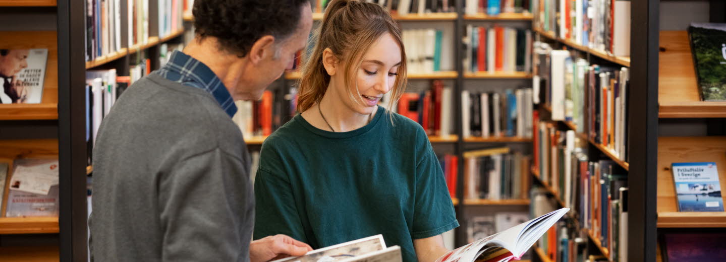 Två bibliotekarier står bland bokhyllor, bläddrar i varsin bok och diskuterar innehållet. 