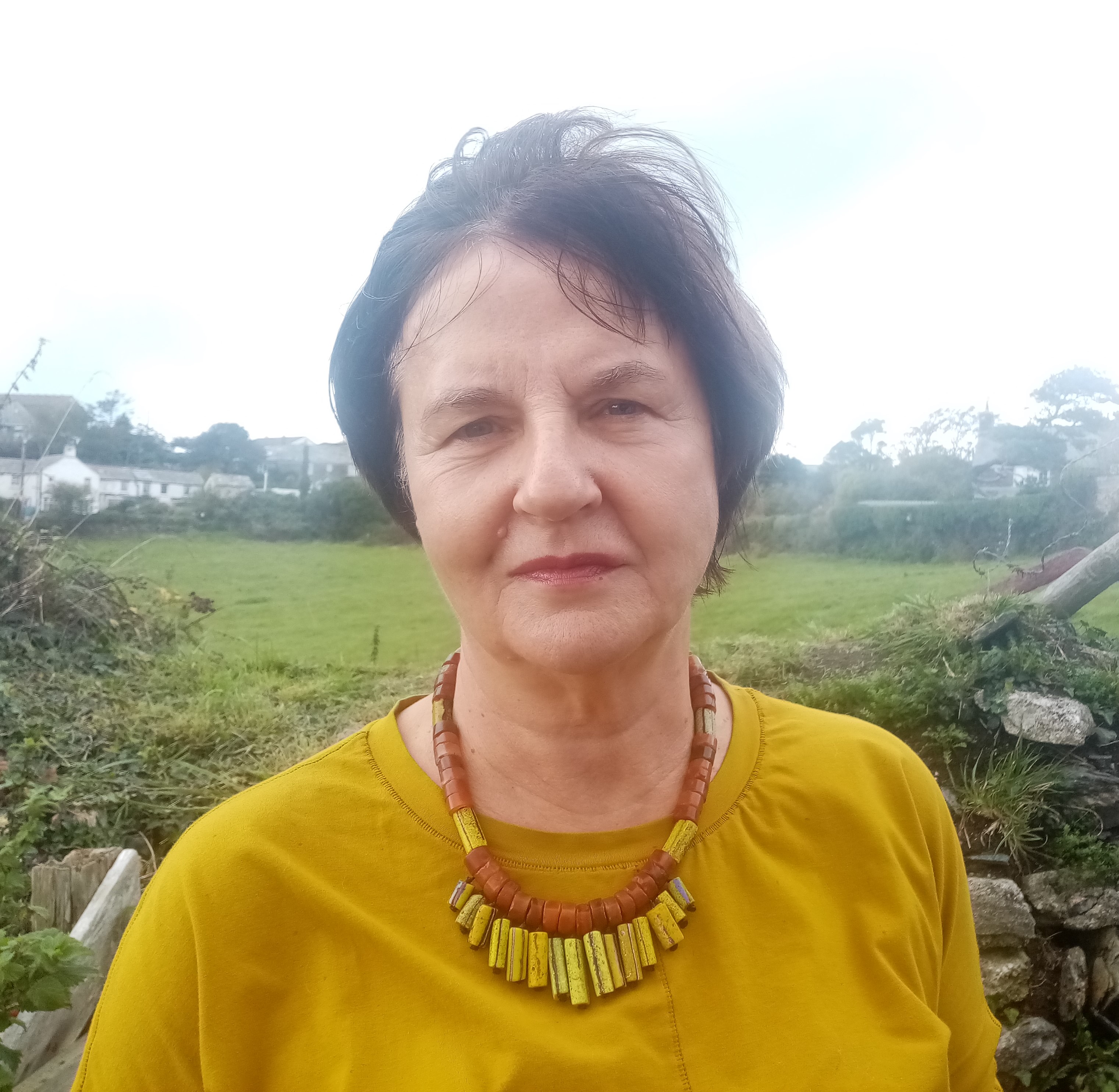 A woman in a yellow top an black short hair is standing in a park and looking towards the camera. 