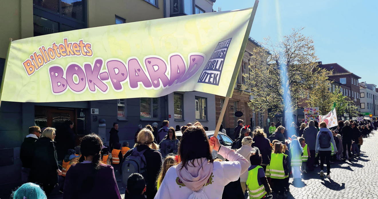Barn och vuxna som tågar längs en gata med banderoller med texten Bibliotekets bokparad.